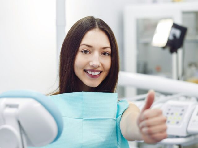 The image shows a young woman with long, dark hair sitting in a dental chair. She is smiling confidently, showing her teeth, and giving a thumbs-up gesture. She is wearing a blue dental bib, indicating that she is in a dental clinic. The background is slightly blurred, with dental equipment and lighting visible, suggesting she has just received or is about to receive dental treatment. The overall mood of the image is positive and reassuring, emphasizing satisfaction with dental care.