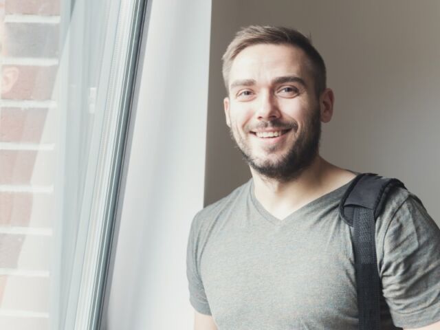 Casual young man with a beard, smiling while standing next to a sunlit window.